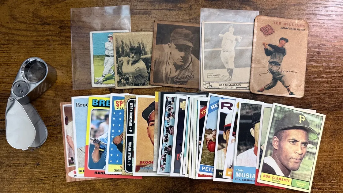 A jeweler's loupe beside two rows of counterfeit baseball cards on a wooden table: a back row of prewar and 1950s fakes including a sleeved Honus Wagner, a Chuck Klein, a Joe DiMaggio and a Ted Williams, and a fanned row of roughly twenty later reproductions in front