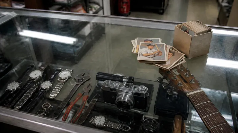 A glass pawn shop display counter holding watches, tools, a camera and a guitar, with a stack of vintage baseball cards and a small box of them resting on top