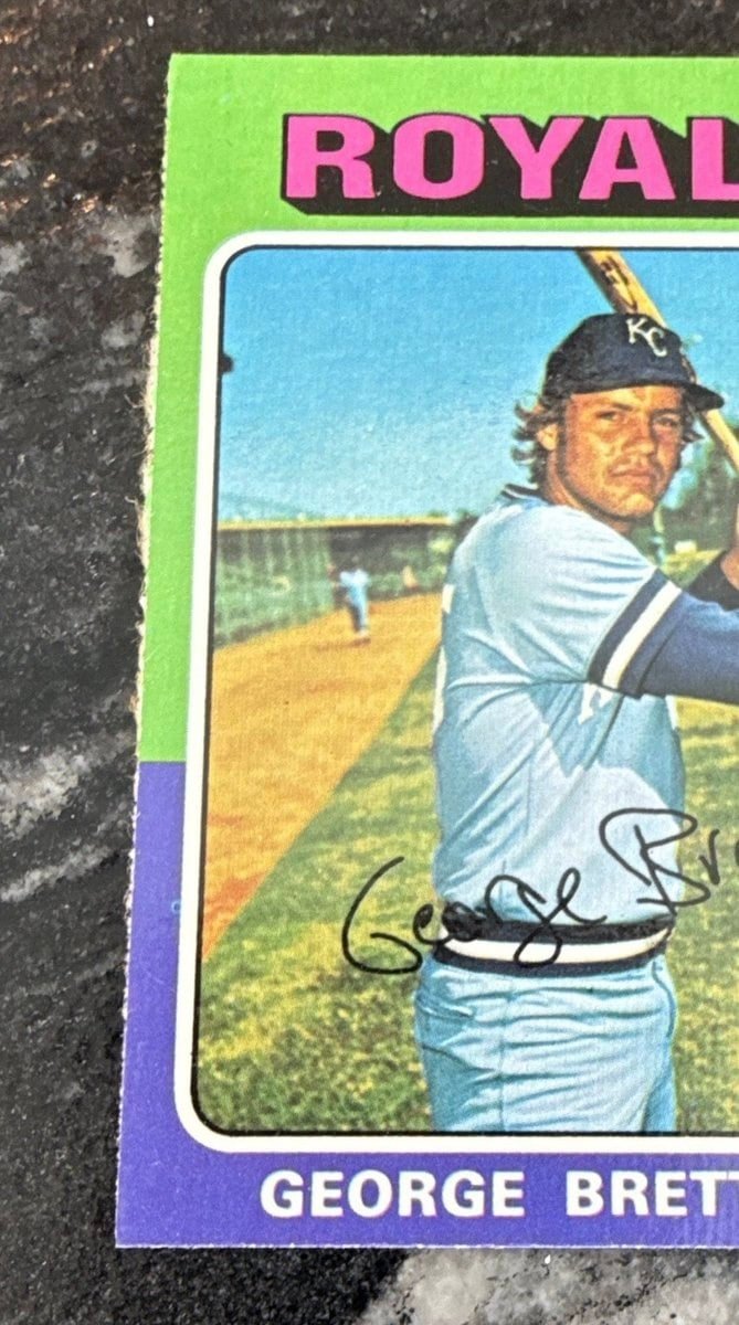 The upper left corner of a 1975 Topps George Brett rookie card, its green and purple border filling the left of the frame. The cut edge is ragged and slightly chewed along its whole length.