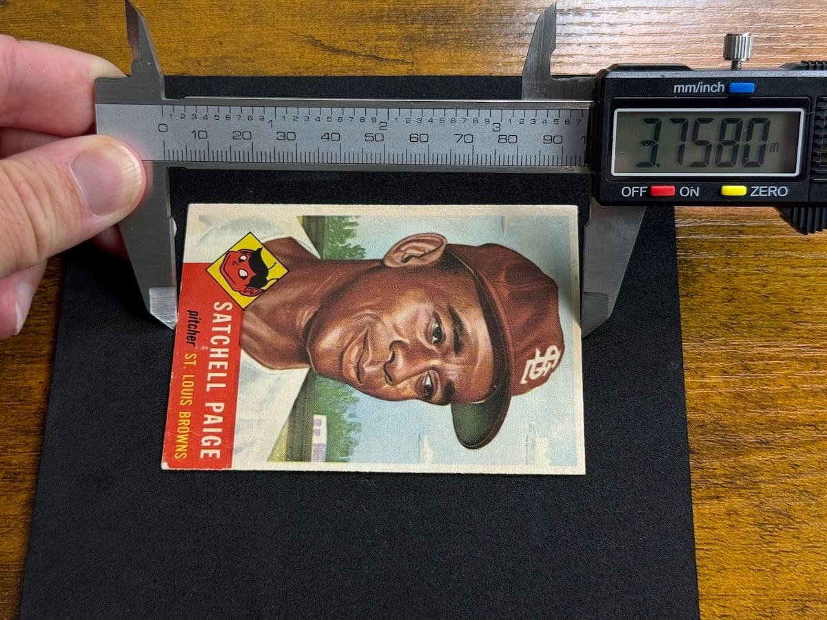 A hand holding digital calipers spanning the long dimension of a 1953 Topps Satchel Paige card lying on a black mat, the display reading 3.7580 inches.